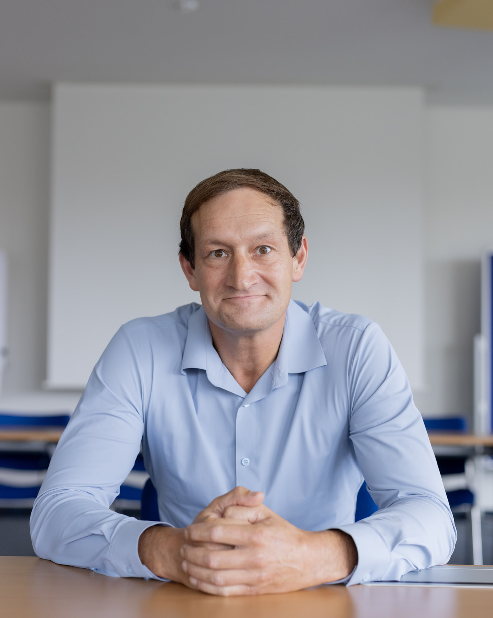 Ein Mann mit kurzem Haar und blauem Hemd sitzt lächelnd an einem Tisch.