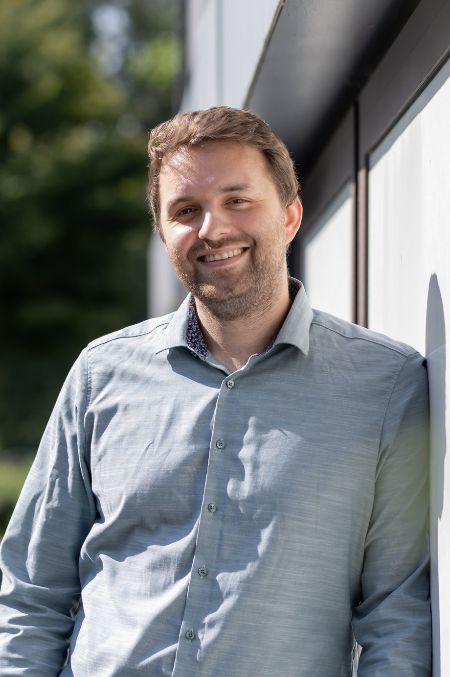 Ein Mann im hellblauen Hemd lehnt lächelnd an einer Hauswand