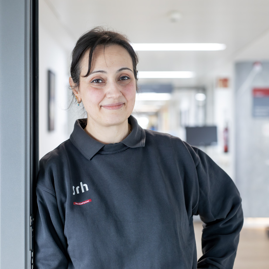 Frau in grauem Polo Shirt steht in einem Krankenhausgang und schaut in die Kamera