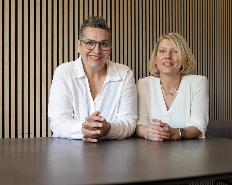 Zwei Frauen in weißen Blusen sitzen an einem Tisch vor einer Holzwand