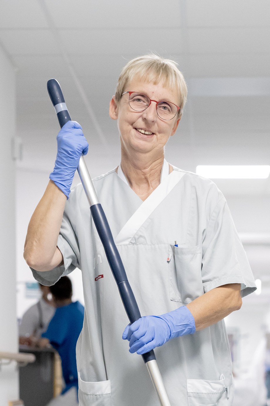 Eine Reinigungskraft hält einen Wischmopp und lächelt in einem Krankenhausflur.