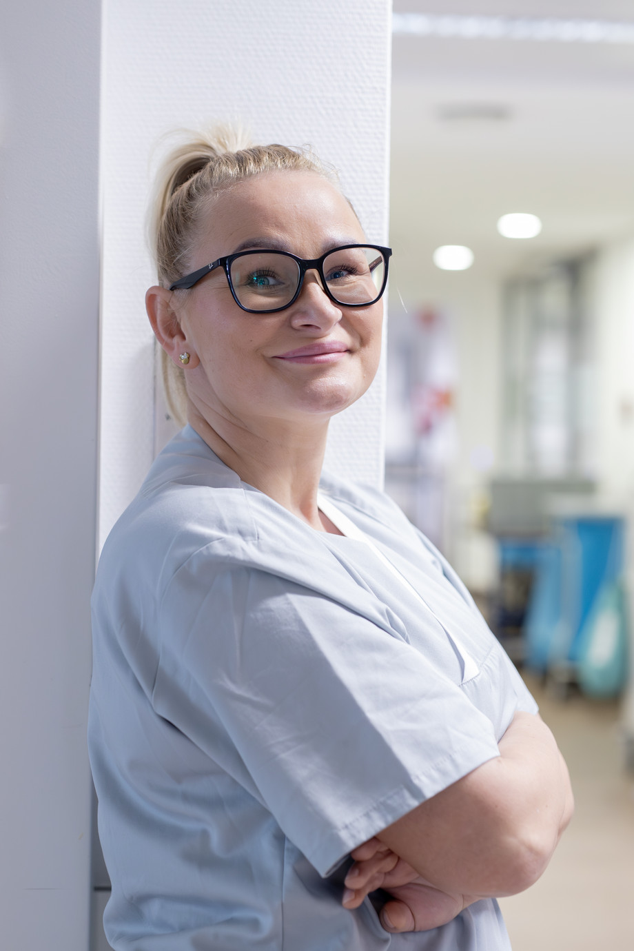 Eine blonde Frau in hellgrauem Oberteil lehnt an einer Wand und schaut in die Kamera