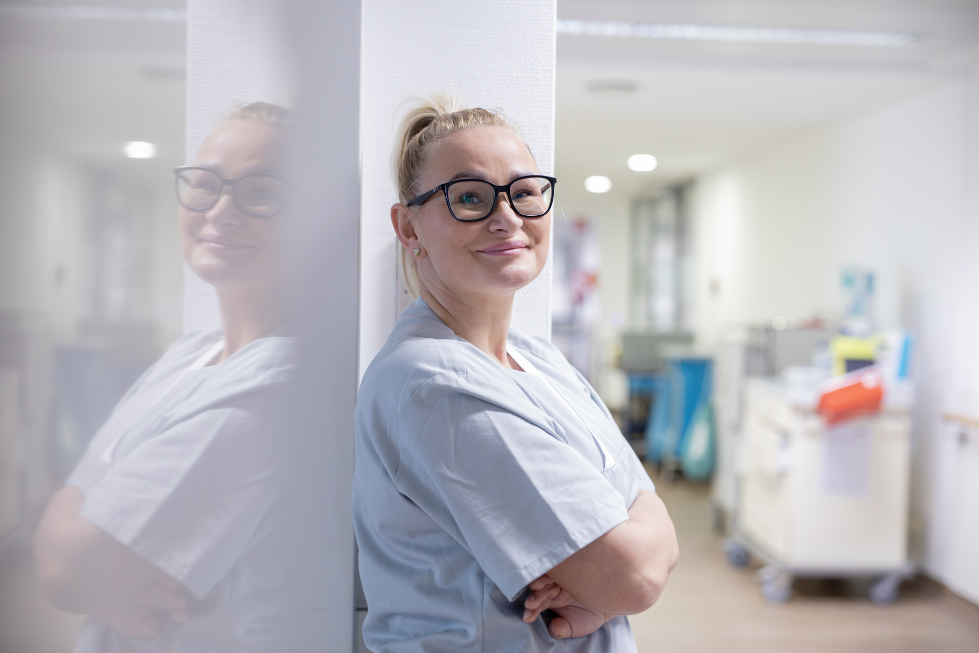Eine blonde Frau in hellgrauem Oberteil lehnt an einer Wand und schaut in die Kamera