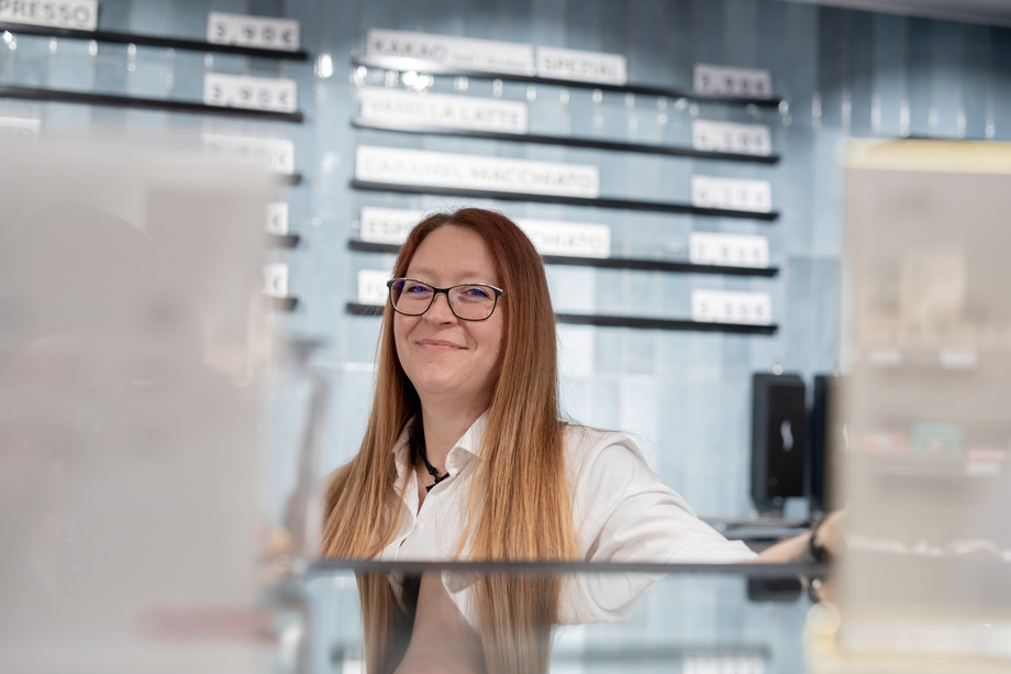 eine Frau mit rotblonden Haaren steht hinter einer Theke und vor einem Preisaushang