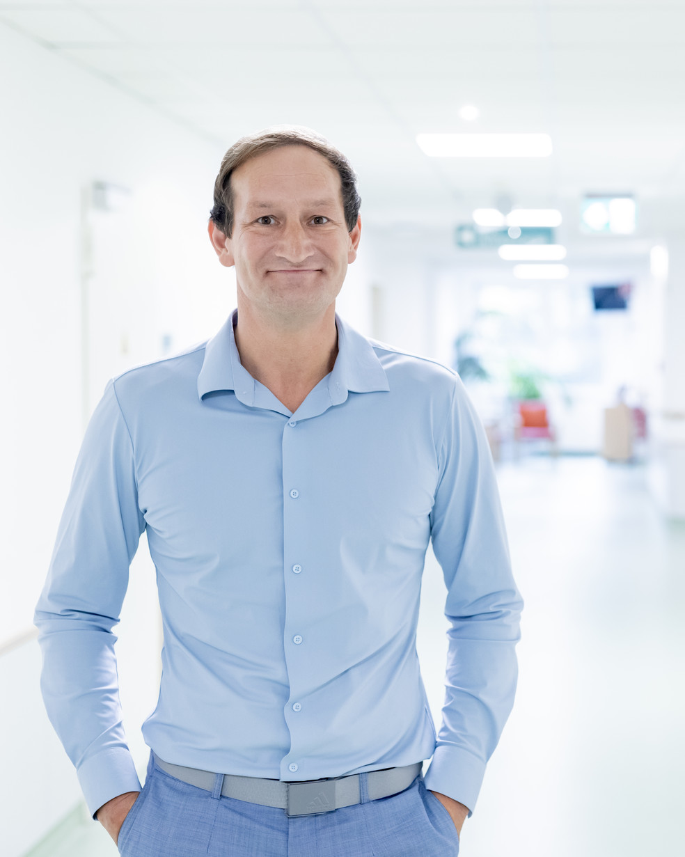 Ein Mann mit kurzem Haar und blauem Hemd steht in einem hellen Flur, mit den Händen in den Hosentaschen.