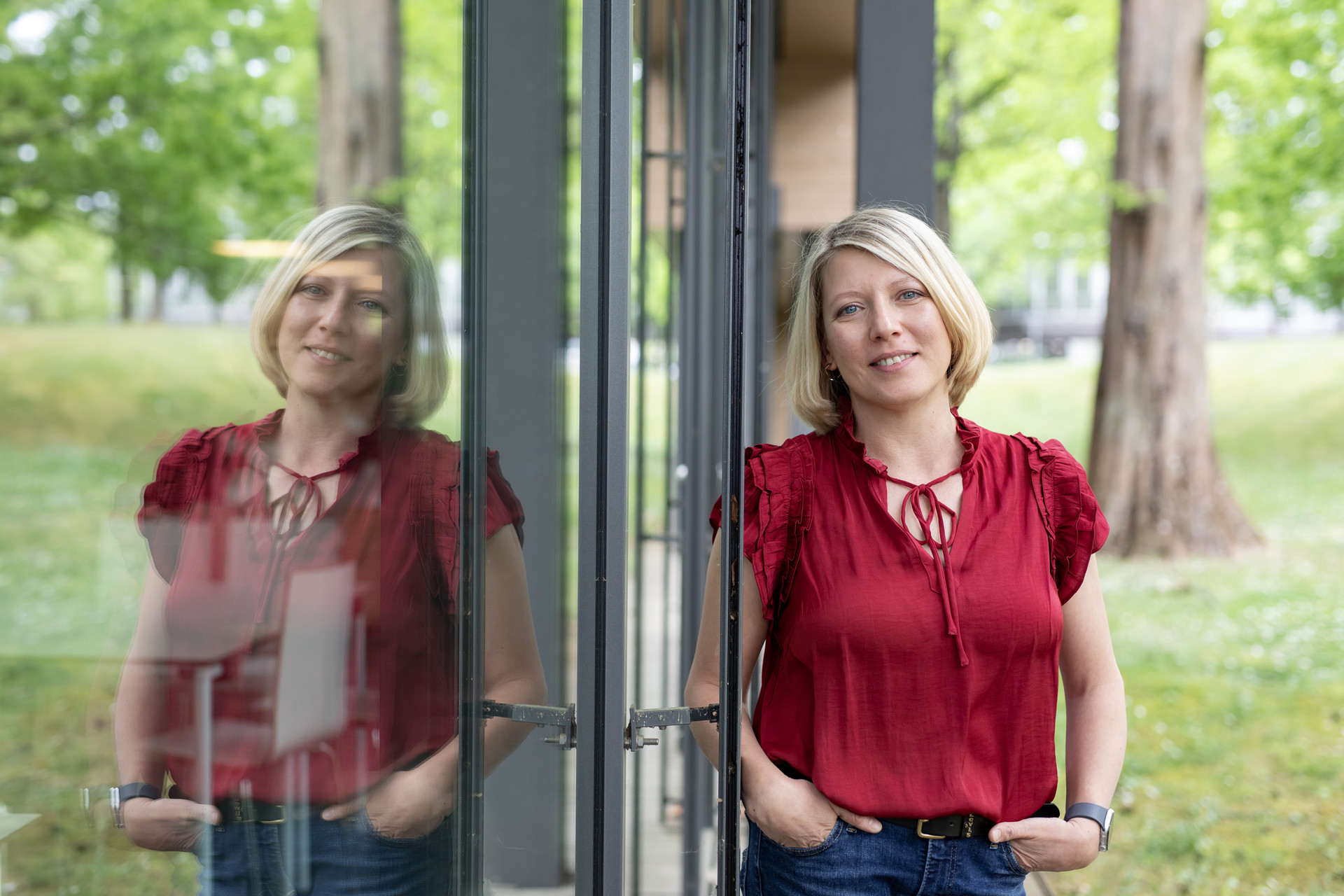 Blonde Frau in roter Bluse lehnt seitlich an einer Glasscheibe