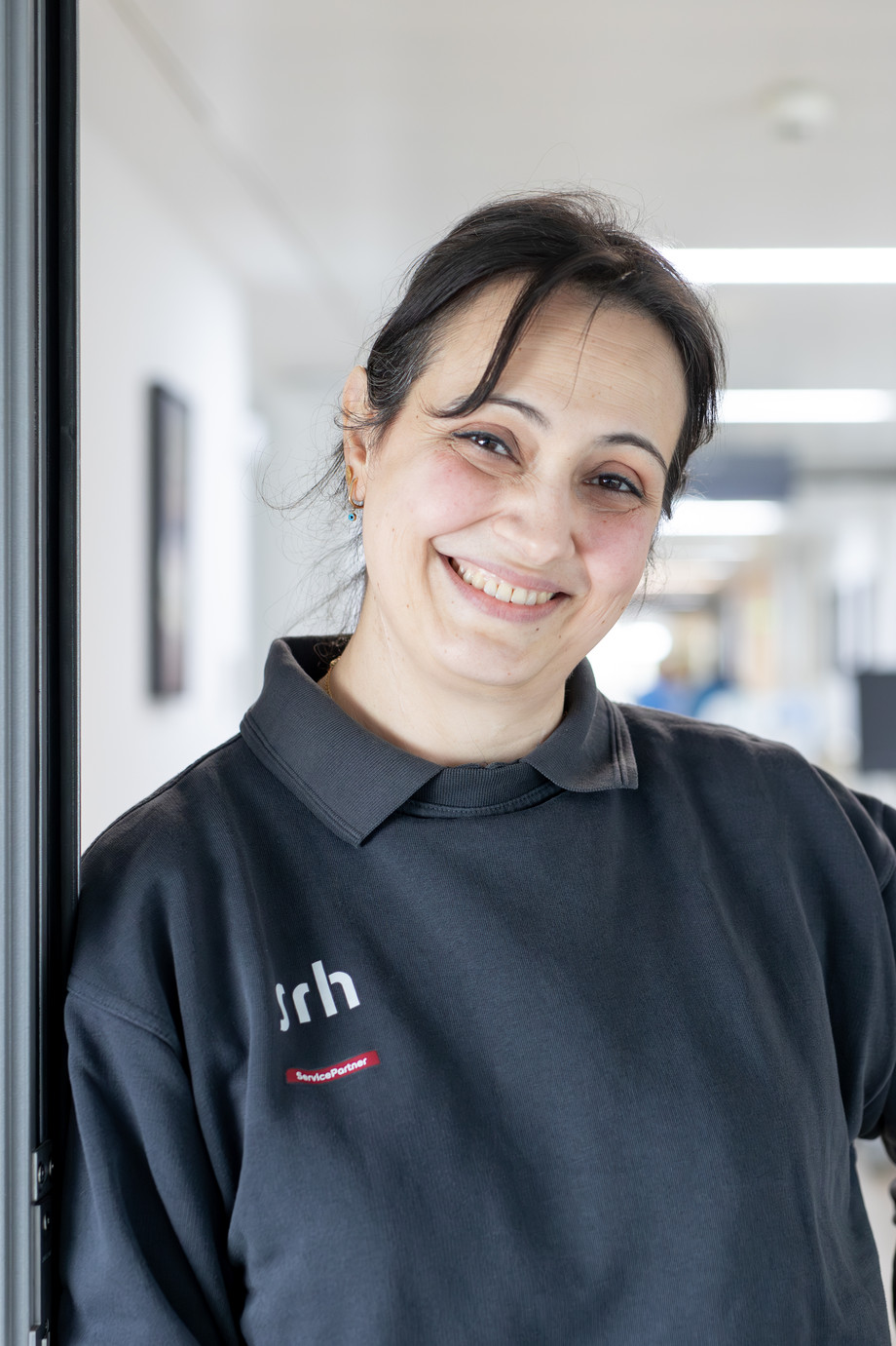 Frau in grauem Polo Shirt steht in einem Krankenhausgang und lächelt in die Kamera