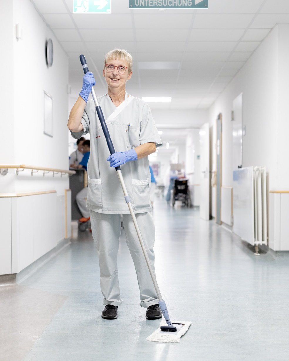 Eine Mitarbeitende aus der Reinigung wischt mit einem Mopp einen Krankenhausflur und hält einen Eimer.